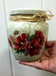 a hand holding a jar with red flowers painted on the side and gold rimmed lid