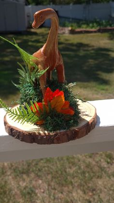 a small toy dinosaur sitting on top of a piece of wood next to some flowers