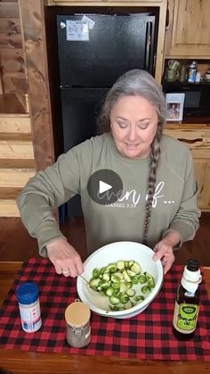 a woman sitting at a kitchen table with a bowl of chopped cucumbers in front of her