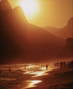 people are walking on the beach as the sun goes down in the mountains behind them