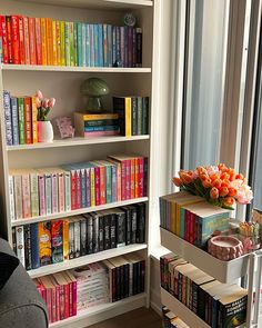 a book shelf filled with lots of books next to a window and a vase full of flowers