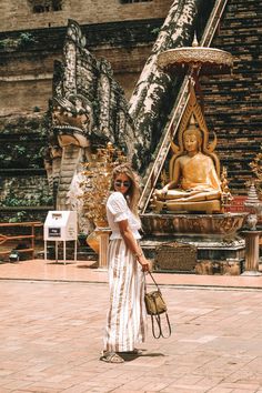 a woman standing in front of a golden buddha statue holding a purse and looking at the camera