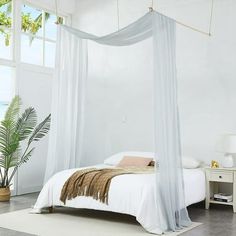 a bed with white sheets and curtains in a room next to a plant on the floor