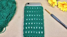 a green crocheted cell phone case next to yellow flowers and a ball of yarn