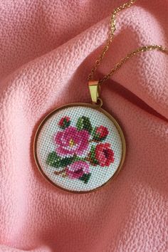 a cross - stitch necklace with pink flowers on it sitting on a pink fabric background