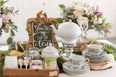 a table topped with dishes and cups filled with flowers next to a sign that says you're home cup of tea