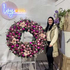 a woman standing in front of a wreath with pink and purple ornaments on it, smiling at the camera