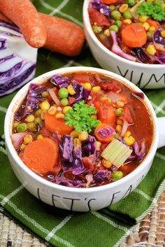 two white bowls filled with vegetable soup on top of a green table cloth next to carrots and cabbage