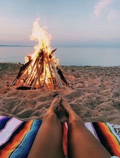 a person sitting in front of a campfire on the beach with their feet up