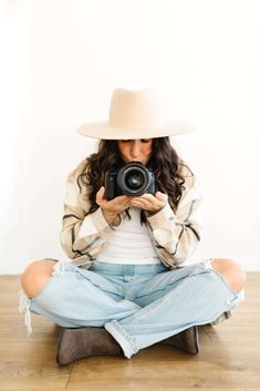 a woman sitting on the floor with her camera in front of her, wearing a hat and jeans