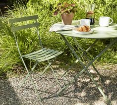 a small table with two chairs and a plate of food on it in front of some bushes