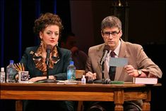 two people sitting at a table with papers and pens in front of them on stage