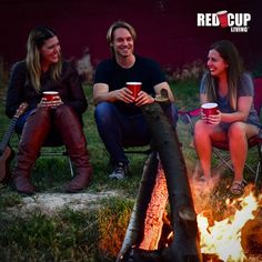 three people sitting around a campfire with cups of coffee in their hands and one holding a guitar