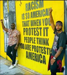 two men standing next to each other near a yellow wall with black writing on it