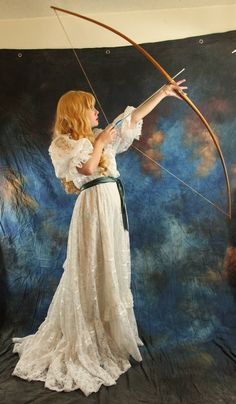 a woman in a white dress is holding a bow and aiming it at the sky