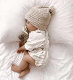 a baby laying on top of a bed next to a teddy bear wearing a hat