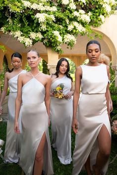 four women in white dresses standing next to each other with flowers on the ground behind them