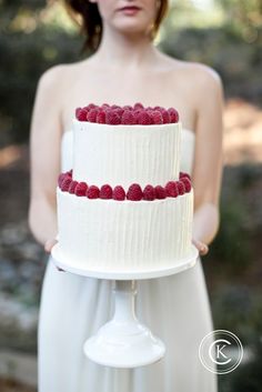 a woman holding a white cake with raspberries on the top and bottom tier