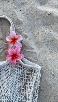 a net bag with pink flowers in it on the beach, next to some sand