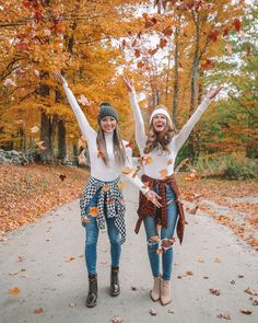 two young women standing in the middle of a leaf filled road with their arms up