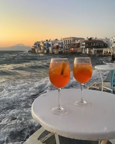 two glasses of wine sitting on top of a table next to the ocean with buildings in the background