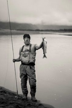 a man holding a fish while standing next to a body of water with a fishing pole