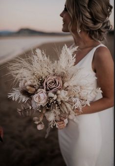 a woman in a white dress holding a bouquet of flowers and feathers on the beach