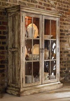 an old china cabinet with glass doors and dishes on it's shelves in front of a brick wall