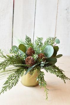 an arrangement of greenery and pine cones sits in a gold vase on a table