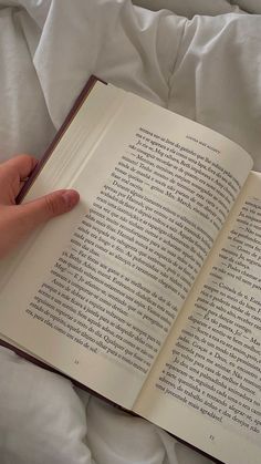 a person is holding an open book in their hand on a white bed spread with sheets