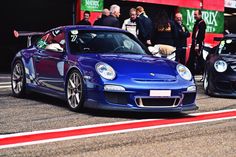 two blue sports cars parked next to each other on a race track with people standing around