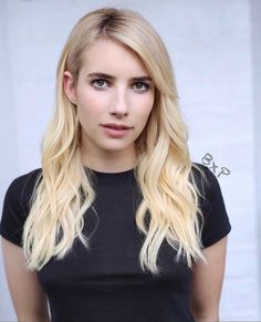 a woman with long blonde hair wearing a black shirt and posing in front of a white wall