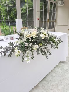 a table with white flowers and greenery on it in front of a large window