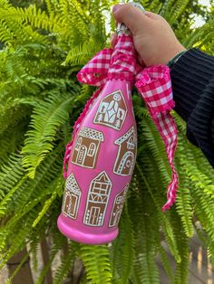 a hand holding a pink and white bottle with houses on it in front of green plants