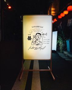 a lit up sign sitting on the side of a road at night with lanterns in the background