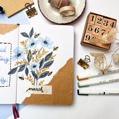 an open notebook with flowers on it next to some markers and pencils in front of them