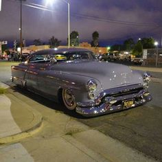 an old car is parked on the side of the road at night with street lights in the background