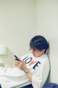 a young man sitting on a bed looking at his cell phone while wearing a white shirt