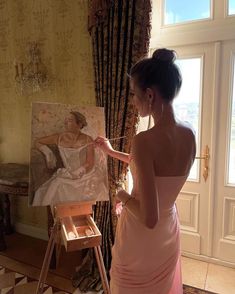 a woman in a pink dress painting on an easel next to a large window