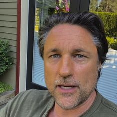 a close up of a person wearing a green shirt near a building and bushes with flowers in the background