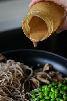 someone pouring peanut butter on noodles in a pan with green peas and mushroom sprouts