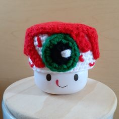 a crocheted red and green hat on top of a white cup sitting on a table