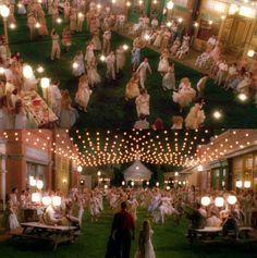 a large group of people are dancing in an outdoor area with lights strung from the ceiling