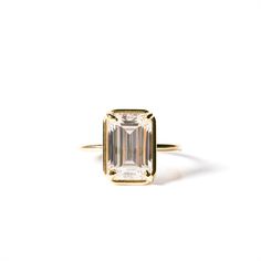 an emerald - cut diamond sits on top of a yellow gold ring, against a white background