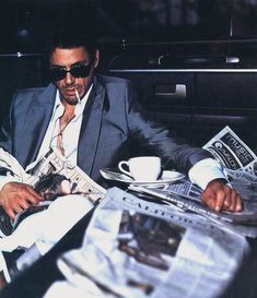 a man in a suit and sunglasses sitting at a table with newspapers