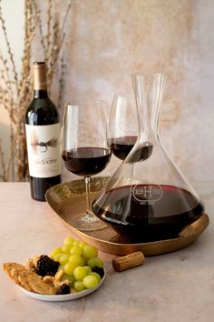 two wine glasses and some grapes on a plate with a bottle of wine in the background