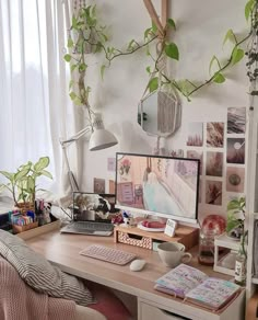 a desk with a computer on top of it next to a window and plants hanging from the ceiling