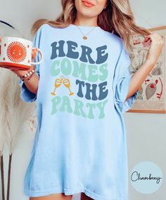 a woman wearing a blue shirt and holding a coffee mug in her right hand with the words here comes the party printed on it