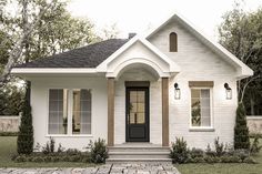 a white brick house with black door and two windows on the front, surrounded by grass and trees