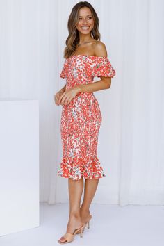 a woman in an orange and white floral print dress standing next to a white backdrop